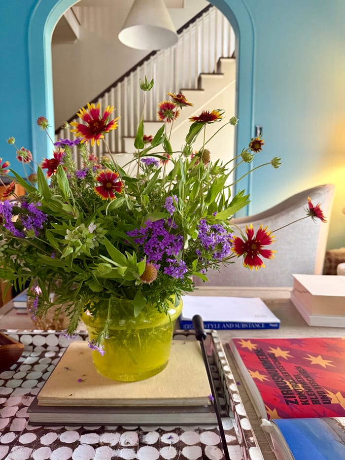 A jar of garden wildflowers on a stack of books, with a pale blue arched mirror behind.