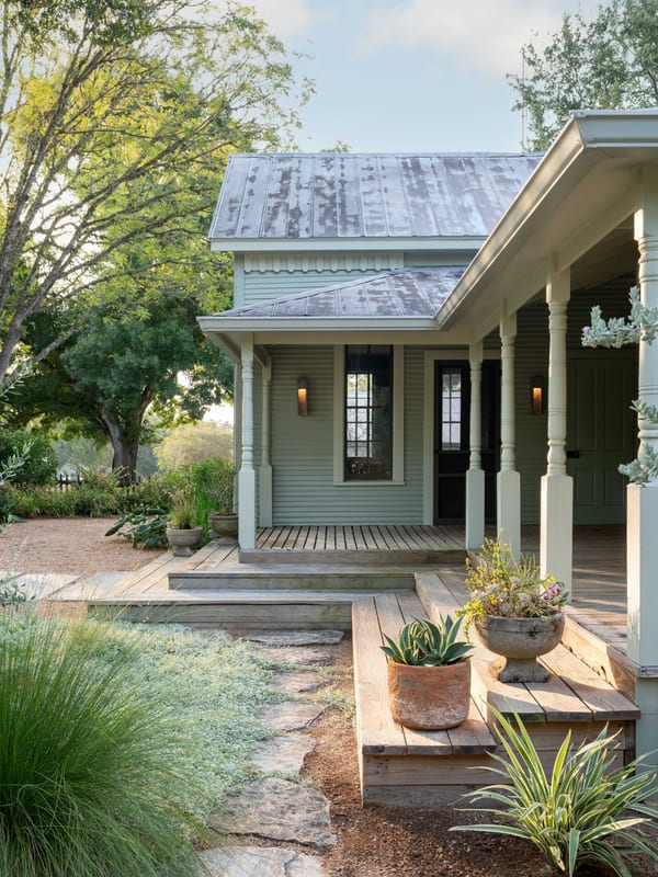 A restored sage-green cottage with a standing-seam metal roof, turned porch posts and a stone path through soft planting.