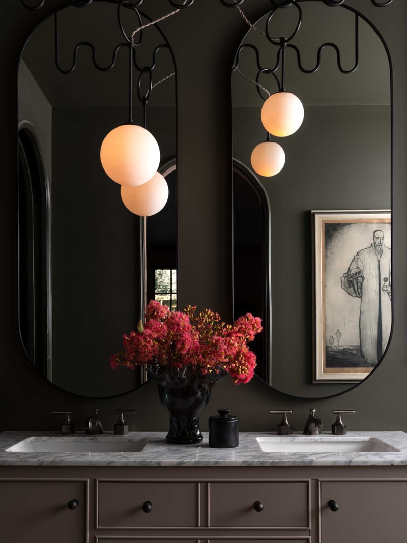 A dark bathroom with twin arched mirrors, globe sconces and red blooms on a marble vanity.