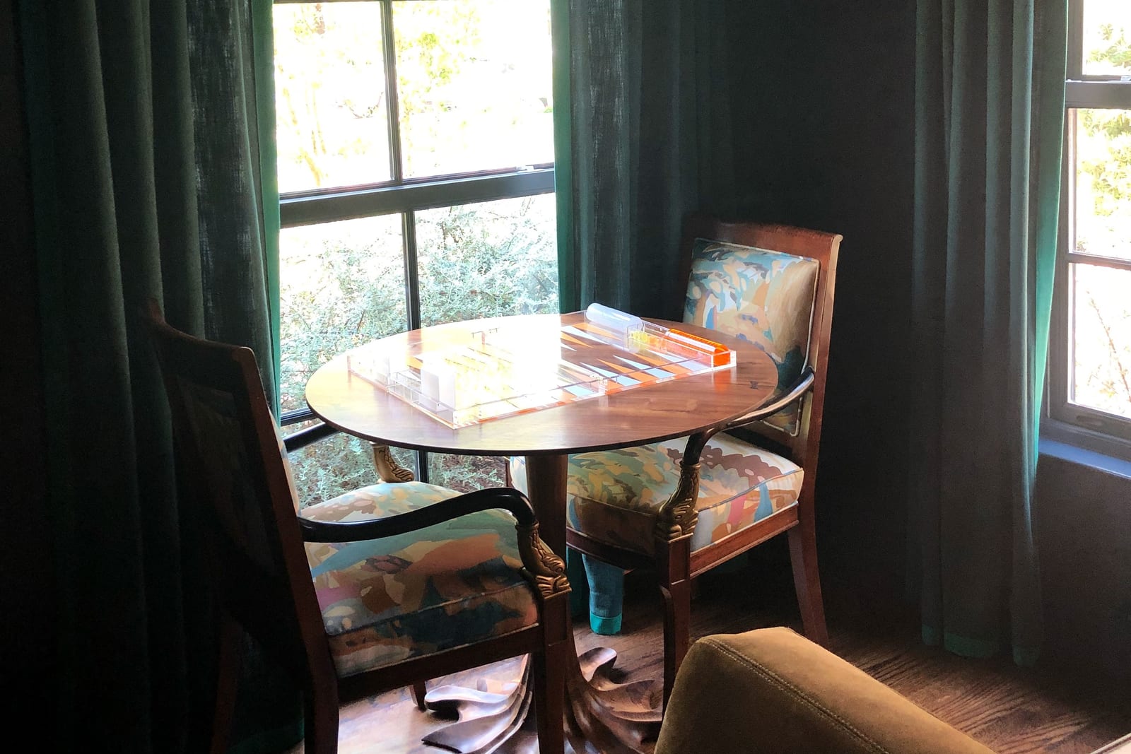 A round game table and two upholstered chairs by a window hung with deep green curtains.