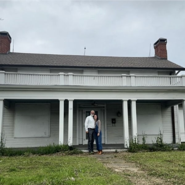 Two people on the porch of the Fox House in 2023, boarded up and before restoration.