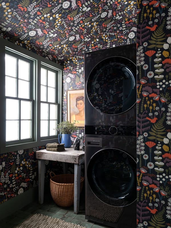 A laundry room wrapped in dark floral wallpaper, with a stacked washer and dryer beside green-trimmed windows.